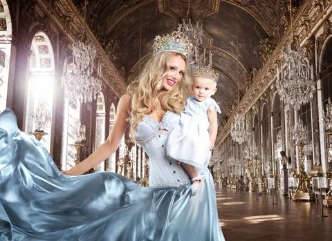 Mother in flowing blue gown holding child in ornate palace corridor setting