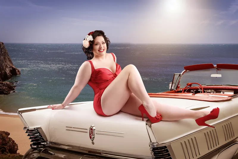Annabel in red vintage-inspired swimsuit reclining on white retro convertible car by dramatic coastal cliffs